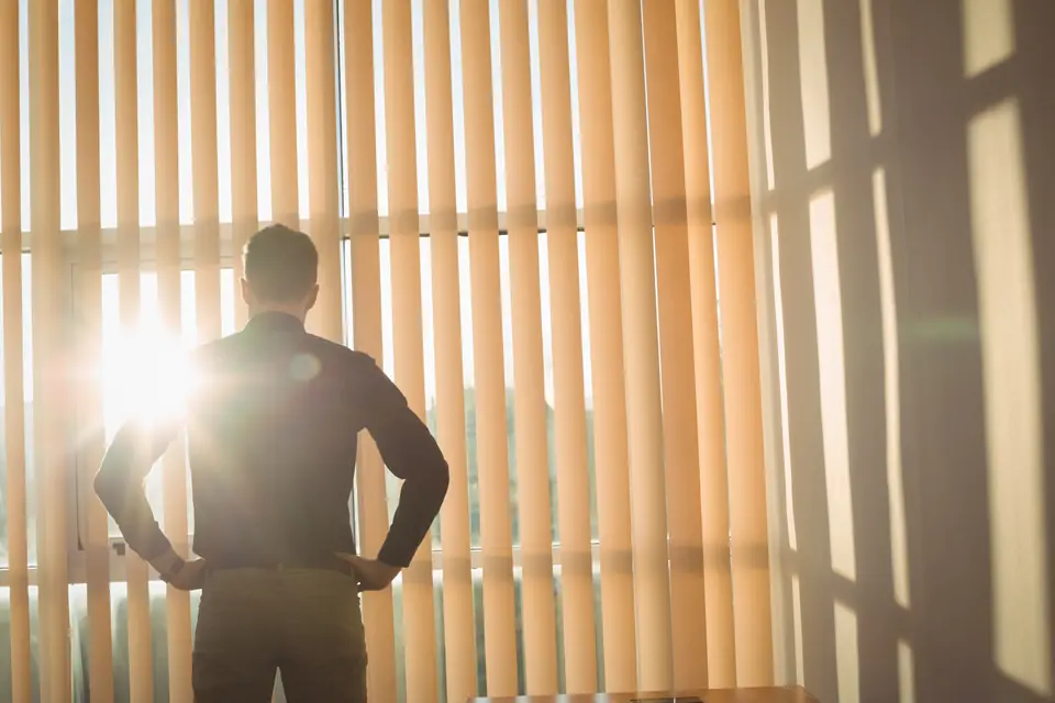Homem de costas olhando pela janela, com o sol entrando entre as persianas.