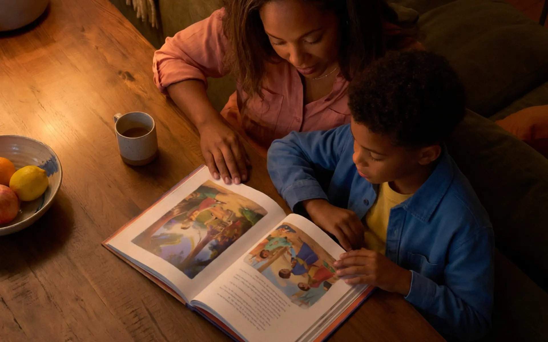 Mãe e filho lendo um livro de figuras na mesa de madeira, à luz da tarde.