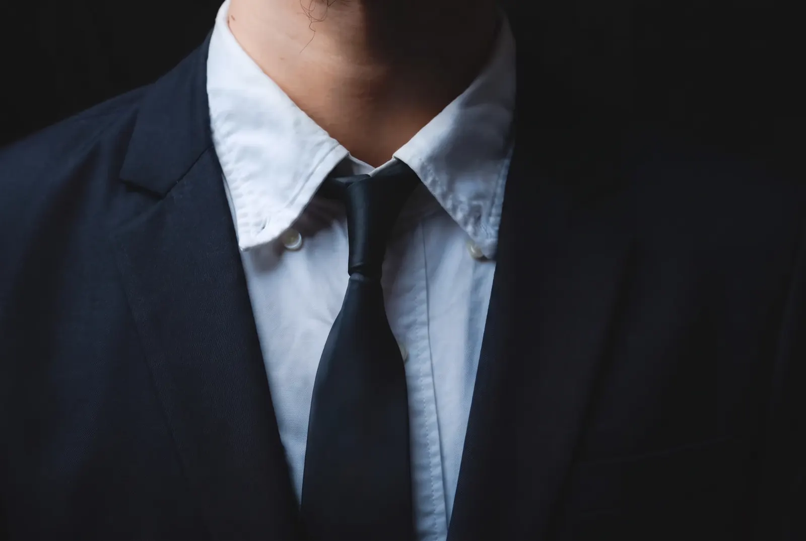 Homem de terno azul-marinho, camisa branca e gravata, sobre fundo escuro.
